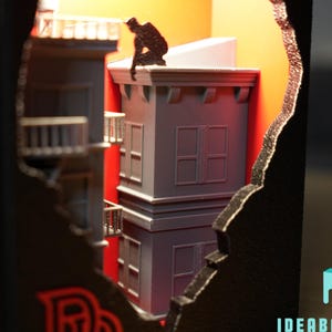 May include: A black shadow box diorama featuring a miniature gray city building with a fire escape and a small dark figure crouching on the roof. The interior is illuminated with a warm orange light, and the text IDEABAYDESIGN is visible.
