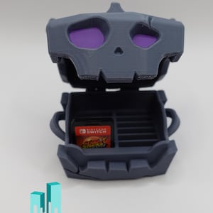 Peut inclure: Un étui en forme de crâne gris avec un motif d'œil violet, ouvert pour révéler un emplacement pour cartouche de jeu Nintendo Switch. L'étui est en plastique imprimé en 3D.