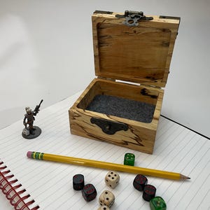 Peut inclure: Une boîte en bois avec un couvercle à charnière et une doublure en feutre gris. La boîte est ouverte et contient plusieurs dés, dont des dés noirs, rouges et verts. Il y a aussi un crayon et une figurine miniature sur un carnet.