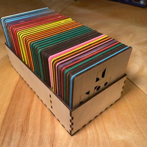 Puede incluir: Una caja de madera llena de piezas de juego rectangulares y coloridas. Cada pieza tiene un borde de diferente color, creando un efecto arcoíris. La caja es de madera clara y tiene diseños recortados.