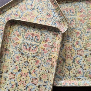 May include: A set of three rectangular metal trays featuring a repeating floral and scrollwork pattern in shades of yellow, pink, blue, and green. Each tray has a raised edge and a smooth surface with a detailed, intricate botanical design.