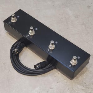 May include: A black rectangular four-button footswitch controller with metallic silver switches labeled 1, 2, 3, and 4. Each switch has a small circular indicator light next to it. A black coiled cable with a multi-pin connector is attached.