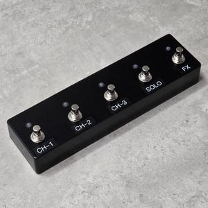 May include: A rectangular black footswitch controller featuring five metallic silver stomp buttons arranged in a row. Each button is labeled with white text reading CH-1, CH-2, CH-3, SOLO, and FX, accompanied by a small circular indicator light.
