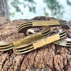 May include: Several antique brass drawer pulls are arranged on a weathered wooden surface. Each pull has a rectangular center and curved, grooved ends. The brass has a vintage, tarnished appearance, suggesting age and use. The background is blurred, with hints of greenery.