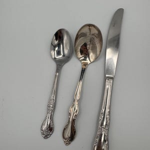 May include: Three pieces of silverware: two spoons and a knife. The spoons have rounded bowls and ornate handles. The knife has a curved blade and a handle with a similar floral design. All pieces are silver-colored and reflect the light.
