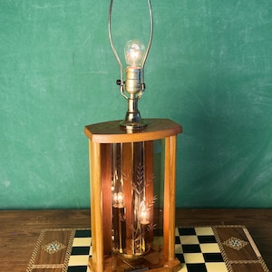 May include: A vintage table lamp with a wooden base and a brass lamp holder. The lamp has a rectangular glass enclosure with etched leaf designs. The lamp is on a wooden surface with a chessboard pattern.