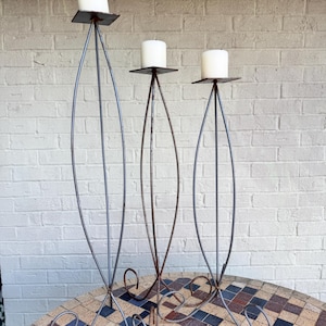 May include: Three tall, slender metal candle holders with a grey finish, each featuring a curved, eye-shaped frame and a square platform at the top. Each holder supports a single white pillar candle, and the bases have decorative curled feet.