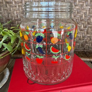 May include: A clear glass jar with a wide mouth, decorated with colorful fruit illustrations. The fruit designs include cherries, watermelon, oranges, and grapes, set against a red grid pattern. The jar is sitting on a red book.