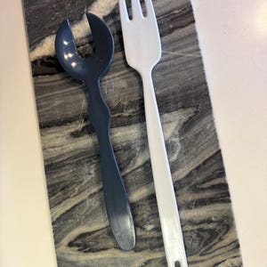 May include: A dark gray salad server and a white salad fork rest on a rectangular marble cutting board. The server has a curved design, while the fork has three tines and a rectangular hole in the handle. Kitchenware.