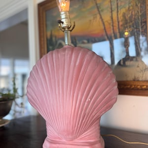 May include: A pink seashell-shaped lamp with a decorative bulb. The lamp has a brass-colored neck and a textured, fan-like design. The lamp is on a dark wooden surface.