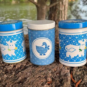 May include: Three vintage blue and white metal canisters with lids. The canisters feature cow and duck illustrations. The canisters are cylindrical and have a blue floral pattern. The lids are blue and the middle canister has a white lid.