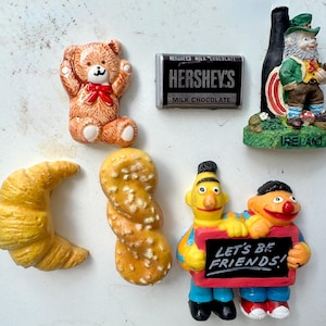 May include: A collection of decorative magnets featuring a teddy bear, a Hershey's chocolate bar, a leprechaun with a bottle, a croissant, a pretzel, and Bert and Ernie holding a sign that says "LET'S BE FRIENDS!"