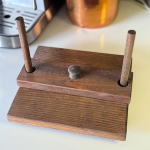 May include: A wooden book press with a dark brown finish. The press has two rectangular wooden platforms, connected by two wooden dowels and a central knob. The wood grain is visible, and the press appears to be handmade.