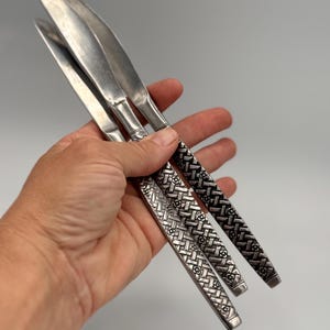 May include: Set of three silver-colored knives with textured handles. The blades are smooth and shiny, while the handles feature a woven floral pattern. The knives are held in a hand against a plain background.
