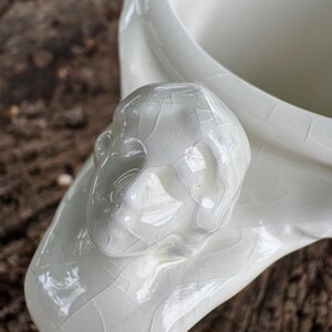 May include: A close-up of a white ceramic vessel with a sculpted human face detail. The vessel has a glossy finish with a cracked glaze effect. The background is a blurred, natural wood surface.
