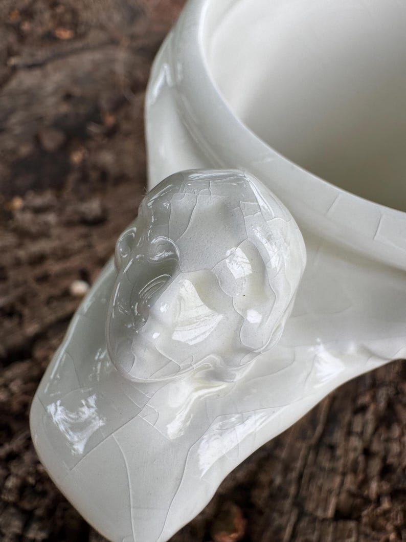 May include: A close-up of a white ceramic vessel with a sculpted human face detail. The vessel has a glossy finish with a cracked glaze effect. The background is a blurred, natural wood surface.
