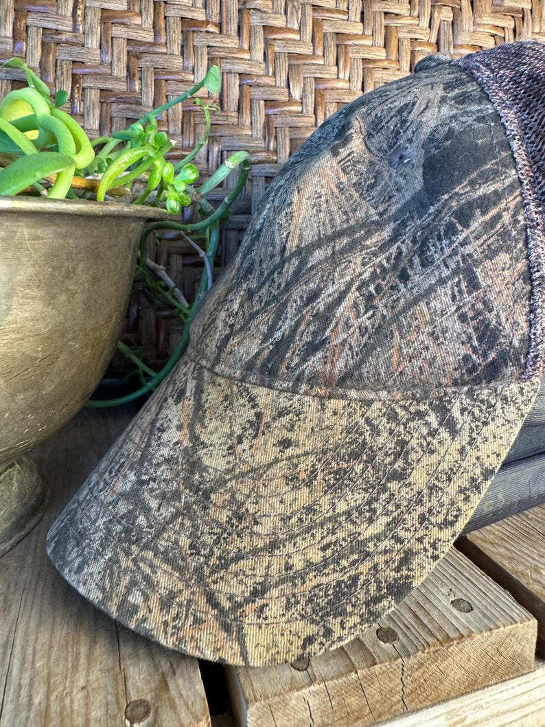 Puede incluir: Gorra de b&eacute;isbol de camuflaje con la parte trasera de malla marr&oacute;n y una visera curva. La gorra presenta un patr&oacute;n de color marr&oacute;n, beige y verde, que se asemeja a un dise&ntilde;o de bosque. El sombrero est&aacute; hecho de una tela duradera.