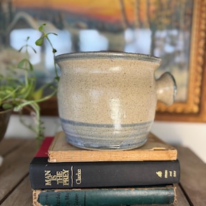 May include: A speckled beige ceramic pot with a blue stripe, featuring a handle. The pot sits atop a stack of vintage books with titles including "Man is the Prey" and "Murder Up Sleeve". The background includes a framed painting.