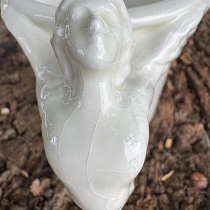 May include: A white ceramic planter with a face sculpted on the side. The planter has a glossy finish and a crackle glaze effect. The face has closed eyes and flowing hair. The planter is likely for indoor use.
