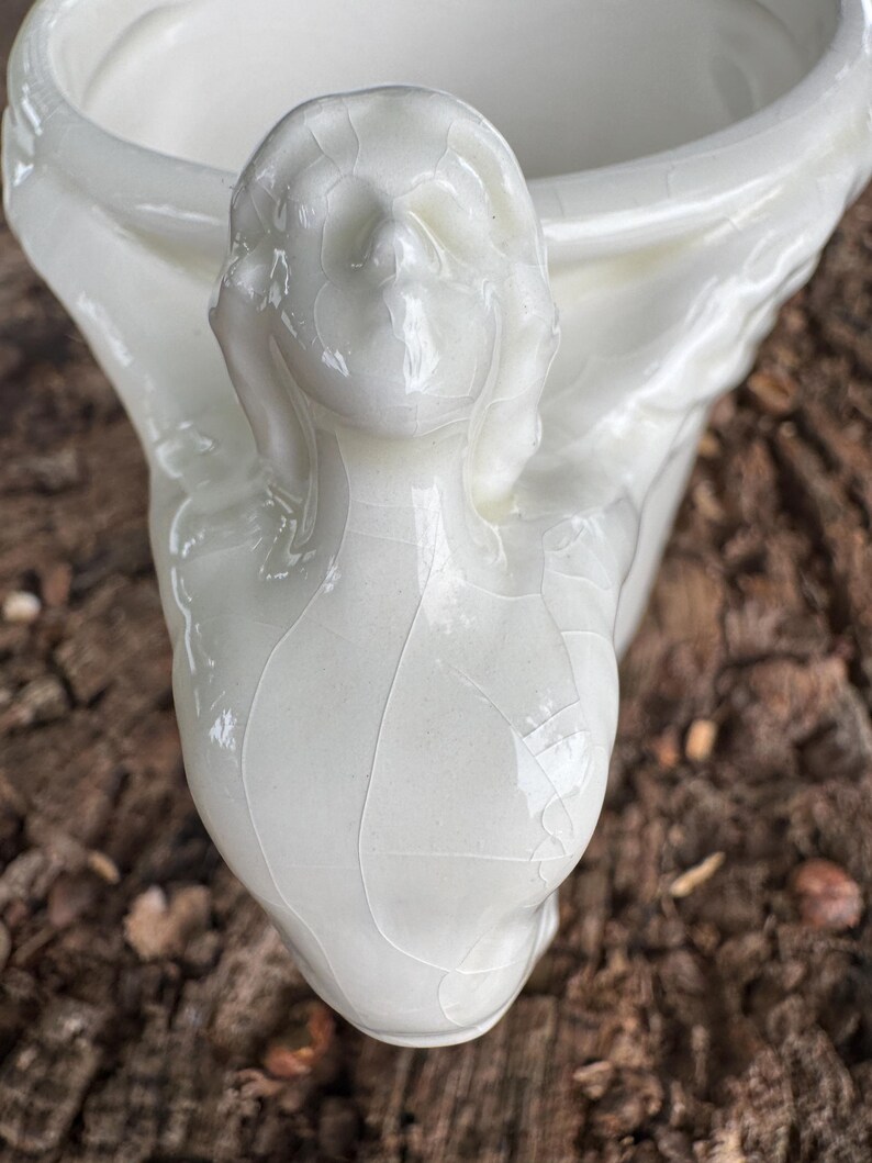 May include: A white ceramic planter with a face sculpted on the side. The planter has a glossy finish and a crackle glaze effect. The face has closed eyes and flowing hair. The planter is likely for indoor use.