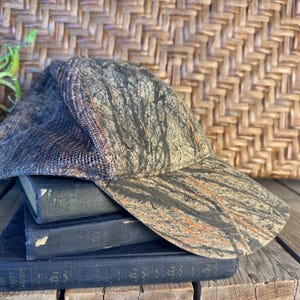 Puede incluir: Una gorra de b&eacute;isbol de camuflaje con la parte trasera de malla descansa sobre una pila de libros antiguos. La gorra presenta un patr&oacute;n marr&oacute;n, negro y naranja. Los libros son de color azul oscuro con letras doradas. El fondo incluye un patr&oacute;n de mimbre tejido y una peque&ntilde;a planta en maceta.
