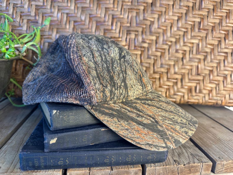 Puede incluir: Una gorra de b&eacute;isbol de camuflaje con la parte trasera de malla descansa sobre una pila de libros antiguos. La gorra presenta un patr&oacute;n marr&oacute;n, negro y naranja. Los libros son de color azul oscuro con letras doradas. El fondo incluye un patr&oacute;n de mimbre tejido y una peque&ntilde;a planta en maceta.