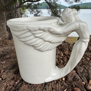 May include: A white ceramic mug with a unique design. The mug features a sculpted angel figure as the handle, with detailed wings extending around the cup. The mug has a slightly cracked glaze, adding to its vintage aesthetic.