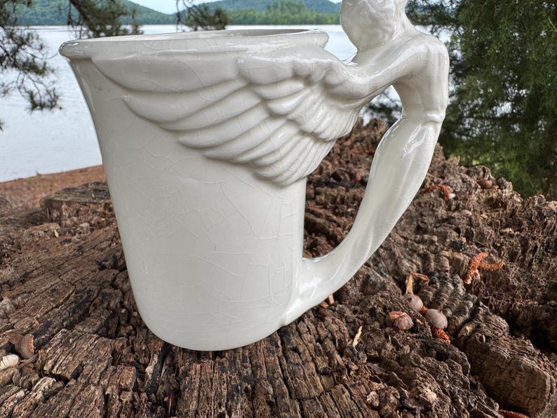 May include: A white ceramic mug with a unique angel wing handle. The mug features a detailed wing design on the side and a sculpted angel figure forming the handle. The mug has a crackle glaze finish, adding to its vintage aesthetic.