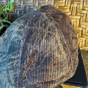 Puede incluir: Una gorra de b&eacute;isbol de camuflaje con la parte trasera de malla. La parte delantera de la gorra presenta un patr&oacute;n de camuflaje verde, marr&oacute;n y negro. La parte trasera es de malla marr&oacute;n. La gorra est&aacute; sobre una superficie de madera.