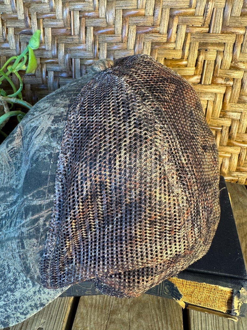 Puede incluir: Una gorra de b&eacute;isbol de camuflaje con la parte trasera de malla. La parte delantera de la gorra presenta un patr&oacute;n de camuflaje verde, marr&oacute;n y negro. La parte trasera es de malla marr&oacute;n. La gorra est&aacute; sobre una superficie de madera.