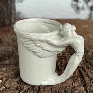 May include: A white ceramic mug with a unique handle shaped like a figure with outstretched arms and wings. The mug has a cylindrical body and a slightly flared rim. The handle is integrated into the design, creating an artistic and functional piece.