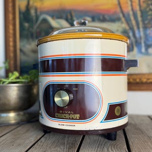 Può includere: Pentola a cottura lenta Rival Crock-Pot vintage in crema, marrone e arancione. L'apparecchio ha un coperchio in vetro e un quadrante con impostazioni basse e alte. La pentola è su una superficie di legno.