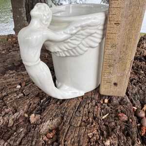 May include: A white ceramic mug with a mermaid handle and wing design. The mug is approximately 4 inches tall, with a detailed mermaid figure forming the handle. The wings are sculpted on the side of the mug, adding a unique touch to this decorative piece.