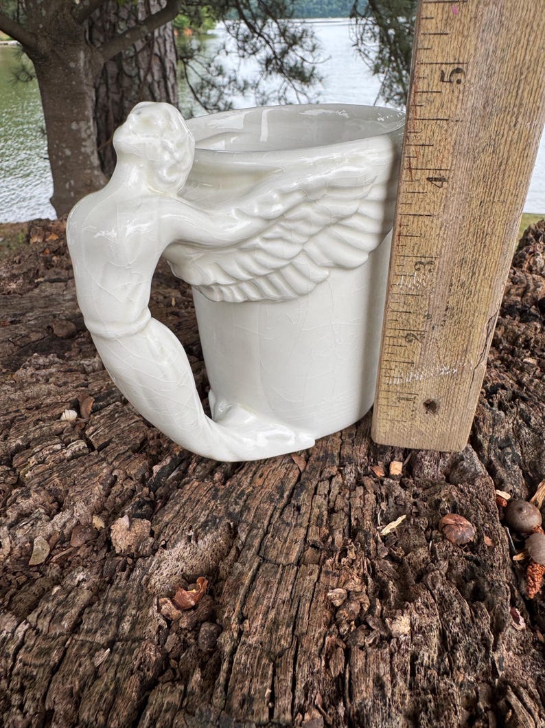 May include: A white ceramic mug with a mermaid handle and wing design. The mug is approximately 4 inches tall, with a detailed mermaid figure forming the handle. The wings are sculpted on the side of the mug, adding a unique touch to this decorative piece.