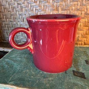 May include: A solid deep red ceramic mug with a smooth, glossy finish and a circular handle. The mug features a slightly flared rim and a cylindrical body, showcasing a consistent monochromatic colour throughout the entire exterior surface.