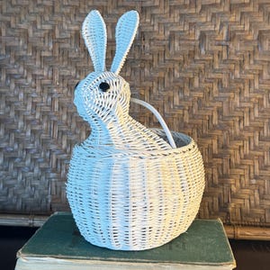 May include: A white woven wicker basket shaped like a rabbit with two tall ears and small black circular eyes. The basket features a rounded body with a tight weave pattern and a curved handle extending from the back of the rabbit figure.