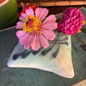 May include: A light blue ceramic pillow-shaped object with a dark blue fern-like pattern and a crackled glaze finish. Two vibrant flowers rest on top: one large pink zinnia with a yellow center and one smaller, dense magenta flower head.