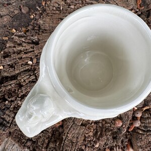 May include: A white ceramic mug with a decorative handle featuring a sculpted face. The mug has a wide opening and a slightly textured surface. The mug is set against a natural wood background.