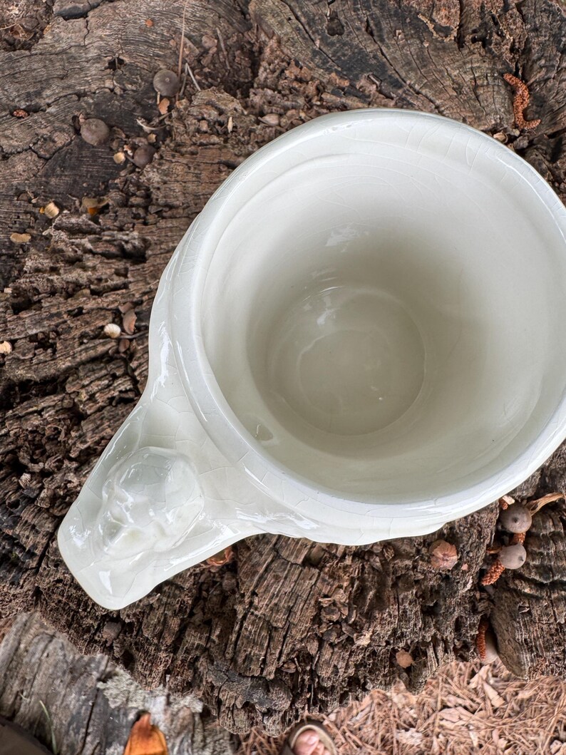 May include: A white ceramic mug with a decorative handle featuring a sculpted face. The mug has a wide opening and a slightly textured surface. The mug is set against a natural wood background.