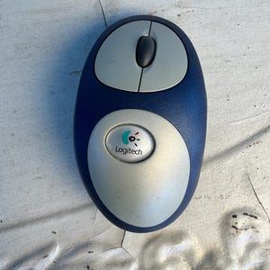 Peut inclure: Une souris d'ordinateur bleu foncé et argentée de forme ovale et arrondie. L'appareil dispose de deux boutons principaux, d'une molette centrale et d'un bouton circulaire argenté au centre affichant le logo Logitech avec une icône colorée.