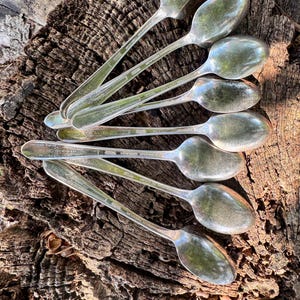 Puede incluir: Una colección de cucharas de plata vintage dispuestas sobre una superficie de madera envejecida y texturizada. Las cucharas tienen un diseño clásico con cuencos alargados y mangos detallados. Las cucharas reflejan el entorno.