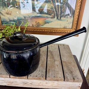 May include: A black enamel saucepan with a long handle and lid, resting on a wooden surface. The pot has a rounded shape and a handle extending to the right. The background includes a framed painting and greenery.