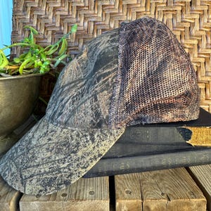 Puede incluir: Gorra de b&eacute;isbol de camuflaje con parte trasera de malla. La gorra presenta un patr&oacute;n marr&oacute;n, verde y negro en la parte delantera y la visera. La parte trasera de malla es de color marr&oacute;n y beige. La gorra descansa sobre una pila de libros viejos y tablas de madera.