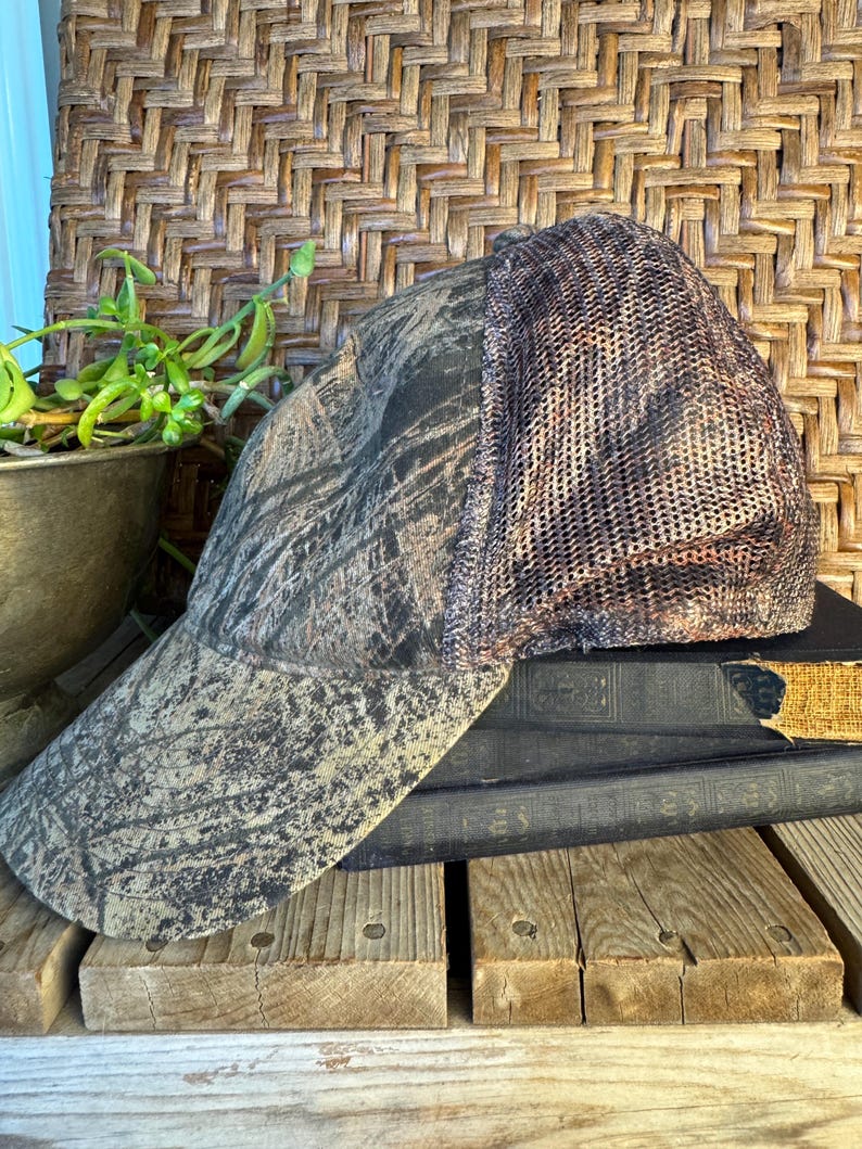 Puede incluir: Gorra de b&eacute;isbol de camuflaje con parte trasera de malla. La gorra presenta un patr&oacute;n marr&oacute;n, verde y negro en la parte delantera y la visera. La parte trasera de malla es de color marr&oacute;n y beige. La gorra descansa sobre una pila de libros viejos y tablas de madera.
