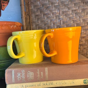 May include: Two ceramic mugs, one bright yellow and one vibrant orange, each featuring a smooth finish, a curved handle, and subtle horizontal ring detailing around the upper body, positioned side by side on top of two stacked hardcover books.