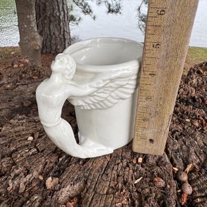 May include: A white ceramic mug with a sculpted angel figure as the handle. The mug features a wing design around the body and is approximately 4 inches tall. The mug is set on a weathered wooden surface.