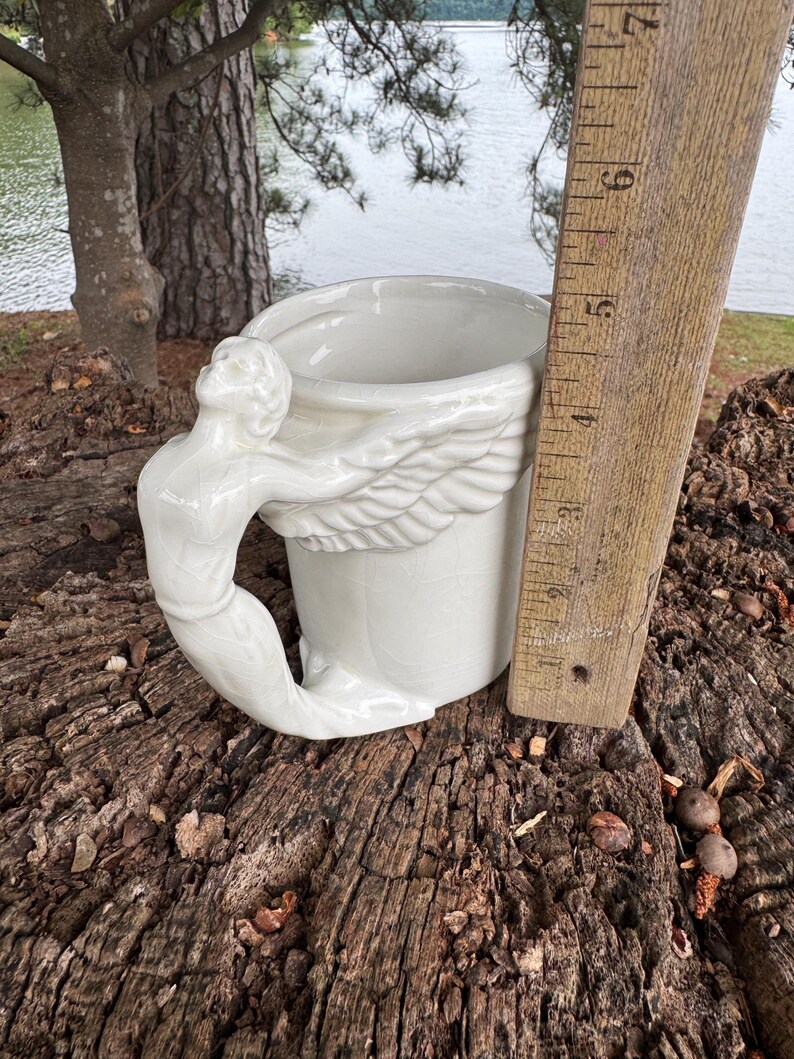 May include: A white ceramic mug with a sculpted angel figure as the handle. The mug features a wing design around the body and is approximately 4 inches tall. The mug is set on a weathered wooden surface.