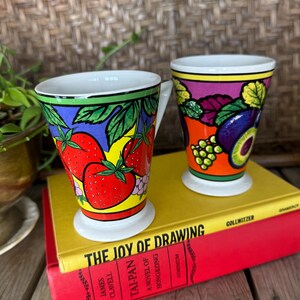May include: Two colorful ceramic mugs with fruit designs. One mug features strawberries, the other has grapes and figs. Both mugs have white bases and handles, and are set on top of two books. The text "THE JOY OF DRAWING" is visible on the yellow book.