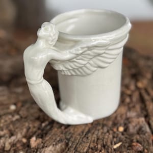 May include: White ceramic mug with a sculpted angel handle and wing detail. The handle is a detailed figure of an angel, and the mug has a smooth, glossy finish. The mug is a unique and decorative item.