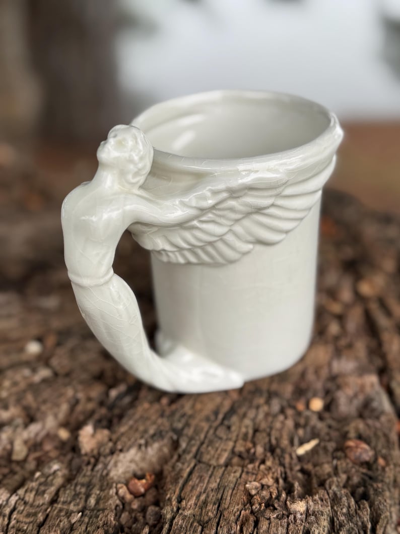 May include: White ceramic mug with a sculpted angel handle and wing detail. The handle is a detailed figure of an angel, and the mug has a smooth, glossy finish. The mug is a unique and decorative item.